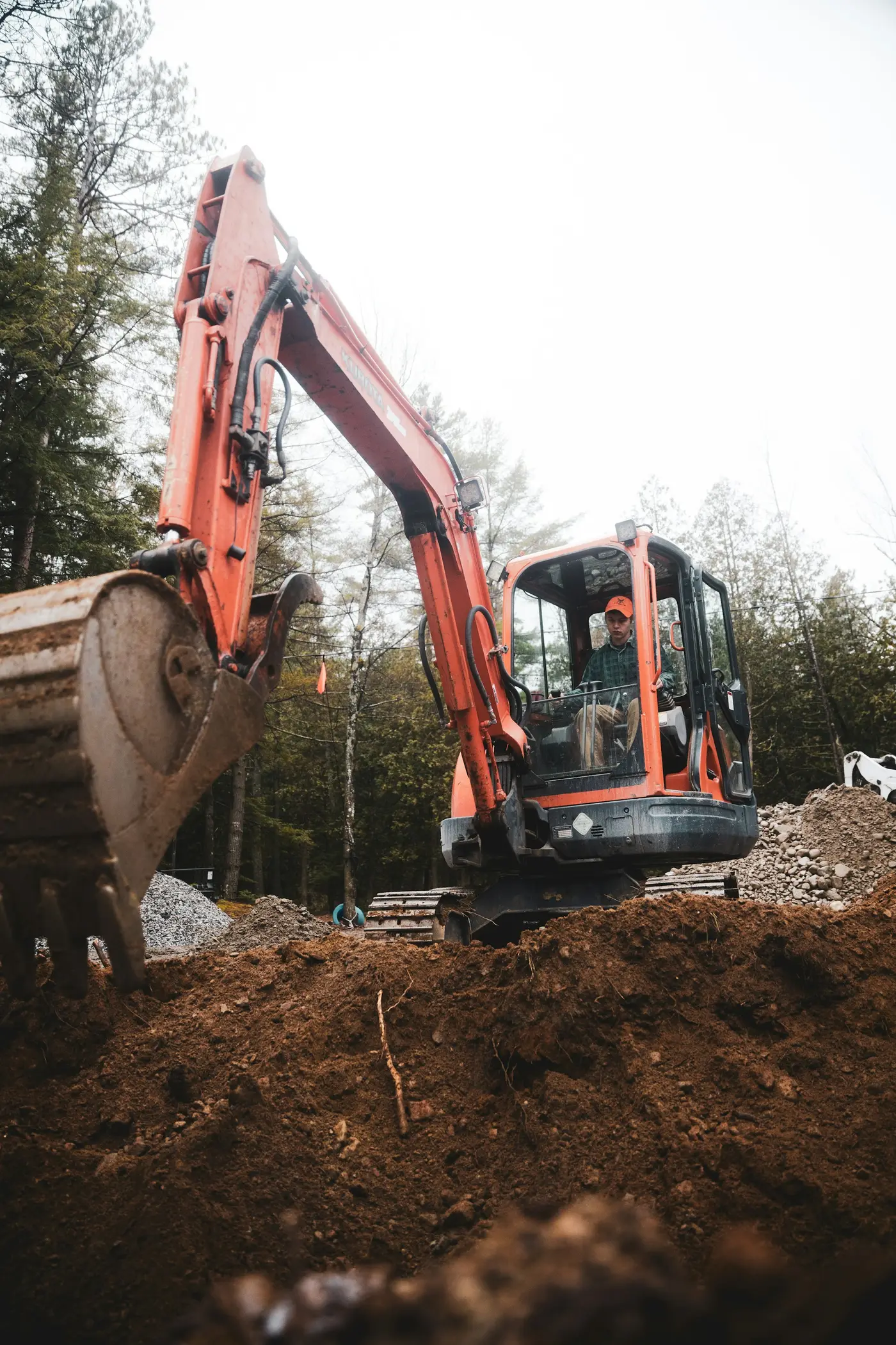 Red compact excavator digging a trench along a house, with the operator in the cab and trees behind