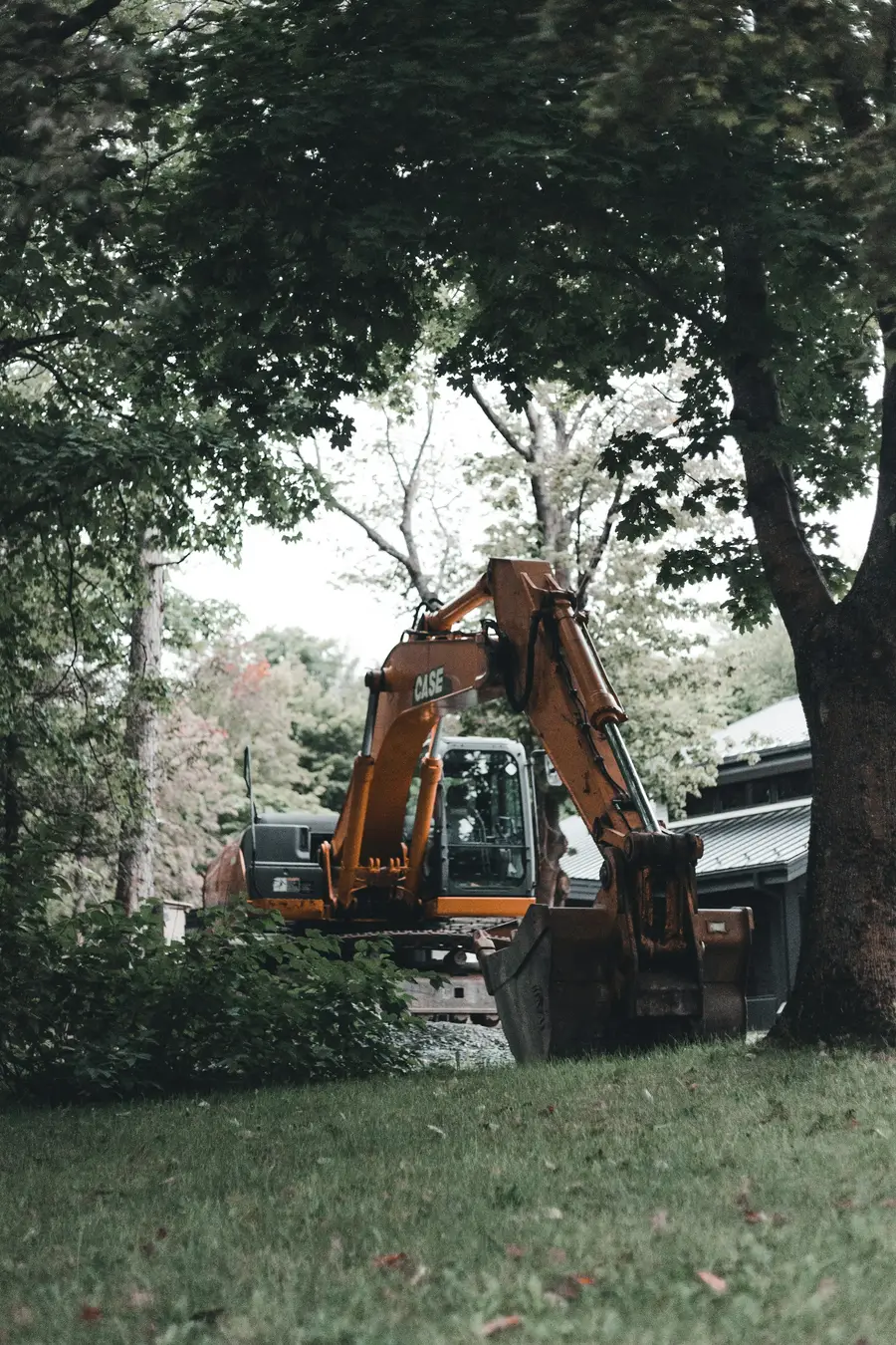 Yellow excavator parked on a lawn under large trees beside a house